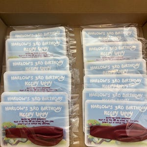 May include: A box of 12 party favors for a 3rd birthday. Each favor is a white card with a blue and white design and the text "Harlow's 3rd Birthday Keepy Uppy". The card has a red balloon attached to it.