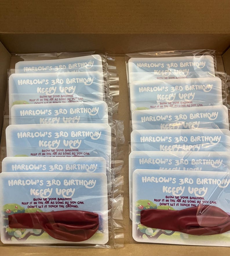 May include: A box of 12 party favors for a 3rd birthday. Each favor is a white card with a blue and white design and the text "Harlow's 3rd Birthday Keepy Uppy". The card has a red balloon attached to it.