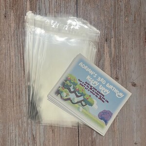 May include: A stack of clear plastic bags with a stack of colorful cards on top. The cards have a cartoon image of a tree with balloons and the text "Adrian's 3rd Birthday Keepy Uppy Blow up your balloon, keep it in the air as long as you can. Don't let it touch the ground."