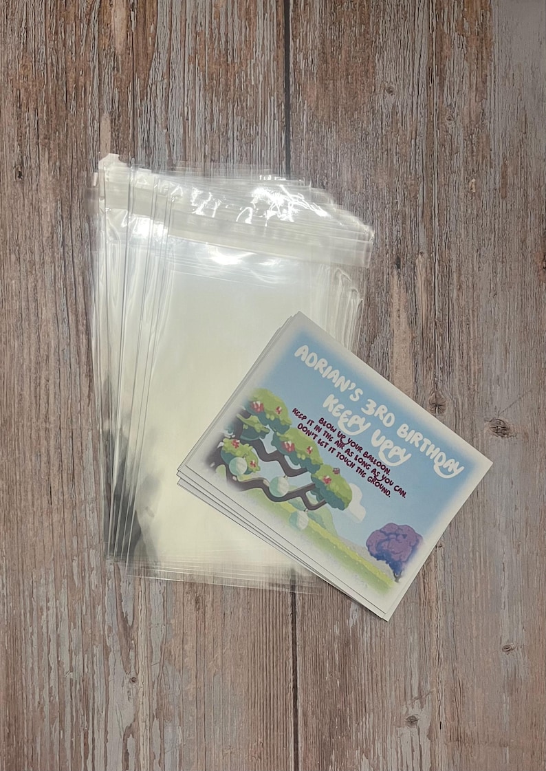 May include: A stack of clear plastic bags with a stack of colorful cards on top. The cards have a cartoon image of a tree with balloons and the text "Adrian's 3rd Birthday Keepy Uppy Blow up your balloon, keep it in the air as long as you can. Don't let it touch the ground."