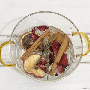 May include: A clear glass bowl with yellow handles filled with water and various botanicals. The bowl contains dried orange slices, cinnamon sticks, rosemary sprigs, apple slices, and red rose petals. The bowl sits on a white wooden surface.
