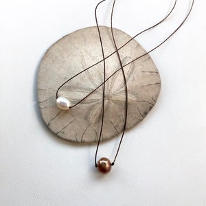 Puede incluir: Dos delicados collares con perlas individuales, una blanca y otra marrón, suspendidas de finos cordones oscuros. Los collares descansan sobre un dólar de arena sobre un fondo blanco, creando una estética minimalista y natural.