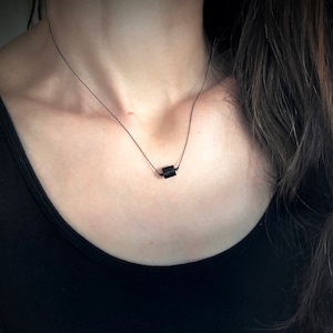May include: A minimalist black necklace featuring a small, rectangular pendant. The pendant is suspended from a delicate black cord, creating a simple yet elegant design. The necklace is worn against a black top.