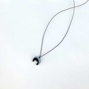 Op de afbeelding: Een minimalistische ketting met een maanvormige hanger. De hanger is zwart en grijs, hangend aan een dun zwart koord. De ketting wordt gepresenteerd tegen een effen witte achtergrond, wat het eenvoudige ontwerp benadrukt.
