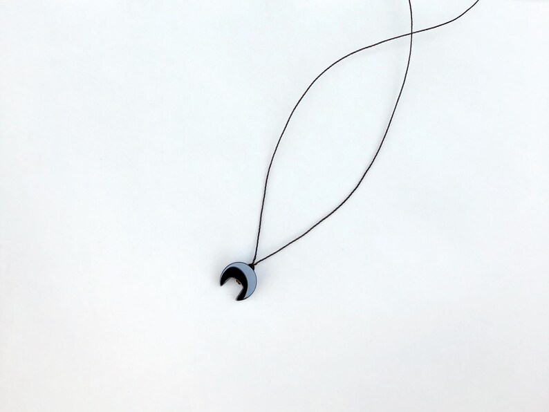 Op de afbeelding: Een minimalistische ketting met een maanvormige hanger. De hanger is zwart en grijs, hangend aan een dun zwart koord. De ketting wordt gepresenteerd tegen een effen witte achtergrond, wat het eenvoudige ontwerp benadrukt.