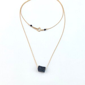 May include: A delicate gold-colored necklace featuring a raw, black tourmaline crystal pendant. The necklace has a double-strand design with a small clasp and black bead accents. The pendant is a rectangular shape.