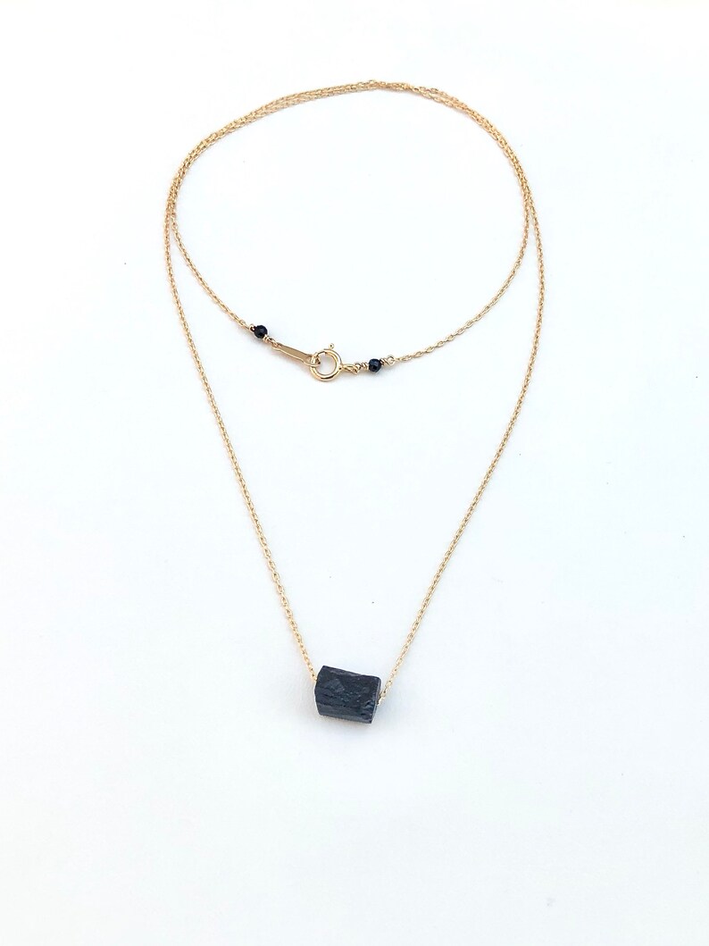 May include: A delicate gold-colored necklace featuring a raw, black tourmaline crystal pendant. The necklace has a double-strand design with a small clasp and black bead accents. The pendant is a rectangular shape.