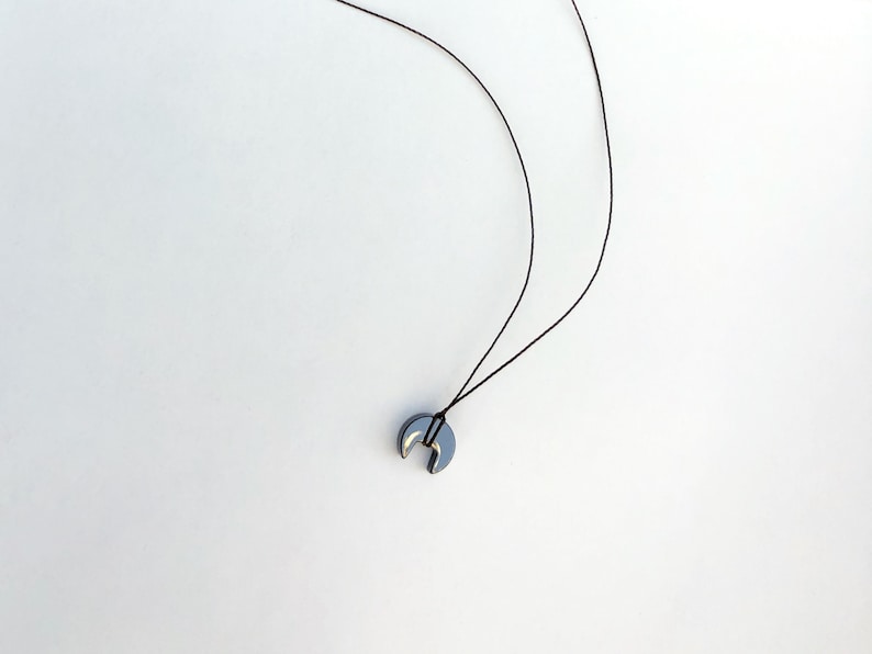 Op de afbeelding: Een minimalistische ketting met een maanvormige hanger in een donkere metallic kleur. De hanger hangt aan een dun, zwart koord. Het ontwerp is eenvoudig en elegant, geschikt voor dagelijks gebruik.