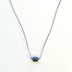 May include: A minimalist necklace featuring a small, dark, square-shaped pendant suspended on a thin black cord. The pendant appears to have a textured surface. The necklace is displayed against a plain white background.
