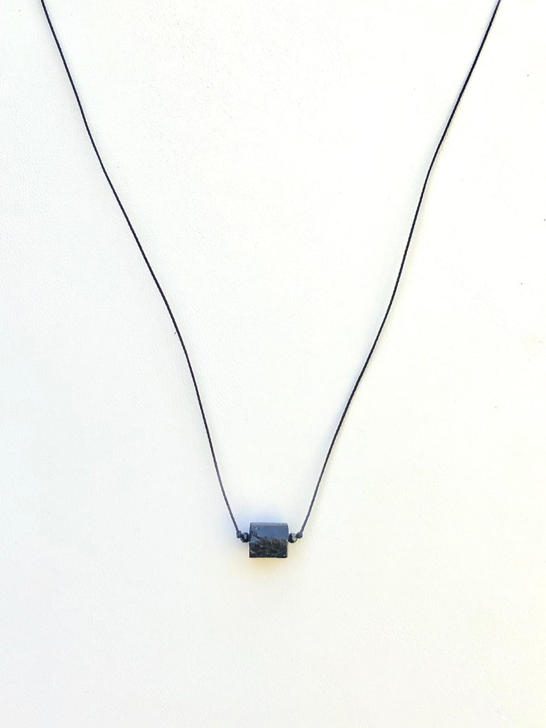 May include: A minimalist necklace featuring a small, dark, square-shaped pendant suspended on a thin black cord. The pendant appears to have a textured surface. The necklace is displayed against a plain white background.