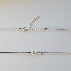 May include: A minimalist necklace featuring a thin black cord with a silver-colored clasp and an adjustable chain. The necklace is displayed against a plain white background, highlighting its simple design.