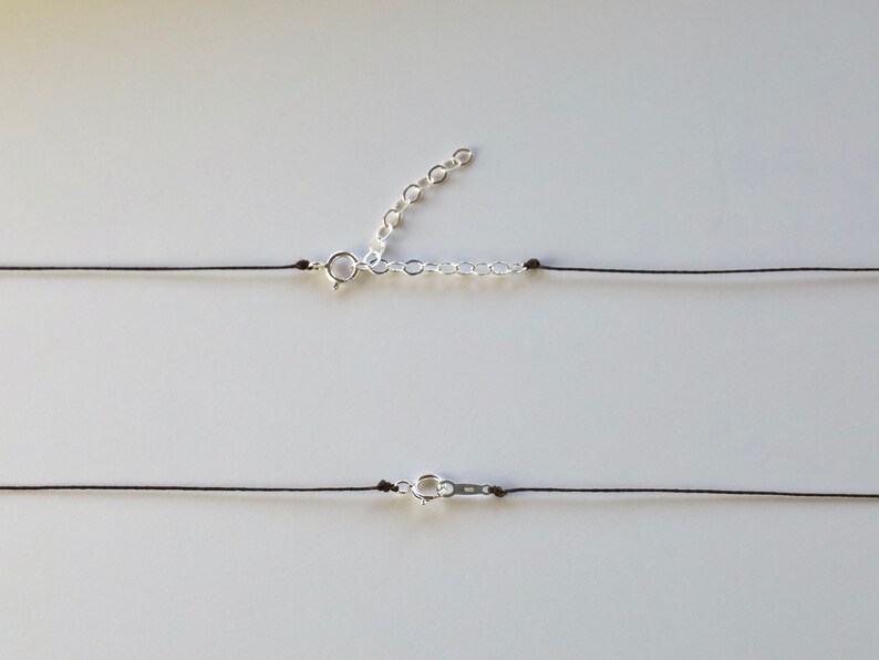 May include: A minimalist necklace featuring a thin black cord with a silver-colored clasp and an adjustable chain. The necklace is displayed against a plain white background, highlighting its simple design.