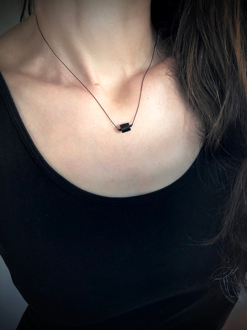 May include: A minimalist black necklace featuring a small, rectangular pendant. The pendant is suspended from a delicate black cord, creating a simple yet elegant design. The necklace is worn against a black top.