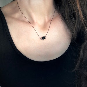 May include: A minimalist black necklace featuring a small, square pendant suspended on a delicate black cord. The necklace is worn against a black top, creating a simple, elegant look. The pendant appears to be made of a solid, dark material.