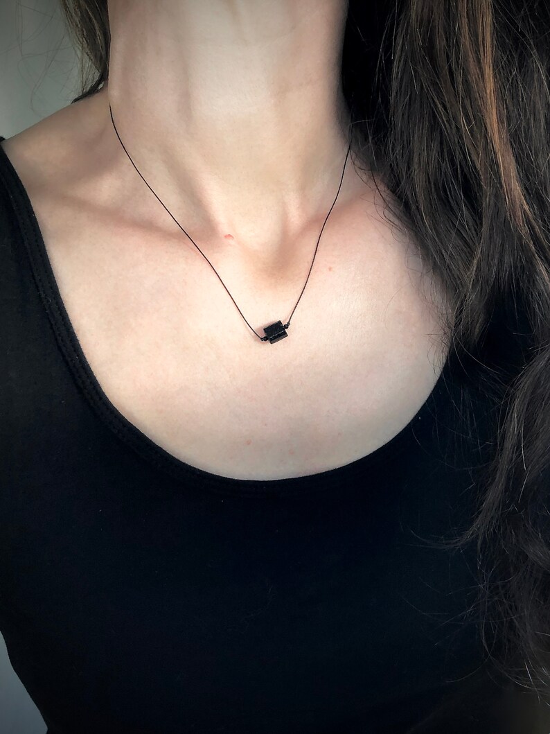 May include: A minimalist black necklace featuring a small, square pendant suspended on a delicate black cord. The necklace is worn against a black top, creating a simple, elegant look. The pendant appears to be made of a solid, dark material.