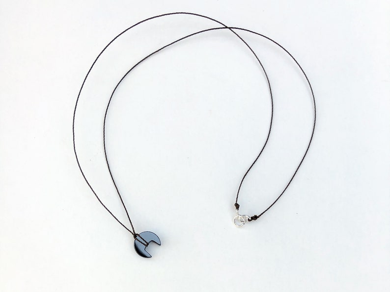 Op de afbeelding: Een minimalistische ketting met een donkergrijze, halvemaanvormige hanger die aan een dun zwart koord hangt. Het koord is vastgemaakt met een zilverkleurige sluiting. De hanger is ongeveer 1,3 cm breed, tegen een witte achtergrond.