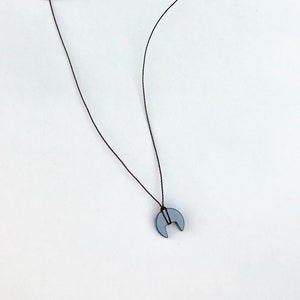 Op de afbeelding: Een minimalistische ketting met een maanvormige hanger in een gedempte grijstint. De hanger hangt aan een dun, zwart koord. De ketting staat tegen een effen witte achtergrond.