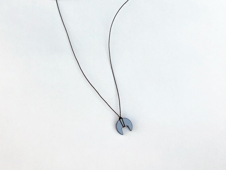 Op de afbeelding: Een minimalistische ketting met een maanvormige hanger in een gedempte grijstint. De hanger hangt aan een dun, zwart koord. De ketting staat tegen een effen witte achtergrond.