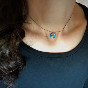 Op de afbeelding: Een delicate ketting met een maanvormige hanger. De hanger heeft een blauwe emaille afwerking met een goudkleurige streep. De ketting wordt gedragen aan een zwart koord, wat een minimalistische en elegante uitstraling cre&euml;ert. De sieraden zijn een modeaccessoire.