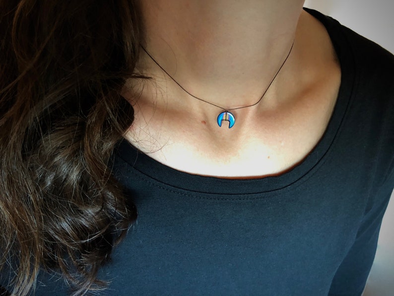 Op de afbeelding: Een delicate ketting met een maanvormige hanger. De hanger heeft een blauwe emaille afwerking met een goudkleurige streep. De ketting wordt gedragen aan een zwart koord, wat een minimalistische en elegante uitstraling cre&euml;ert. De sieraden zijn een modeaccessoire.