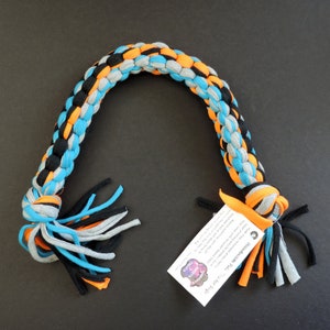 May include: A braided dog toy made from recycled t-shirts in black, gray, orange, and blue. The toy has long, frayed ends. The toy is labeled "Handmade Pet Toy" with a paw print logo.