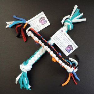 May include: Two handmade dog toys made from braided strips of fabric in white, black, orange, red, and teal. The toys are tied at each end with a knot and have long fabric tails. The toys are labeled "Handmade Pull Toy for Dogs" with a paw print logo.