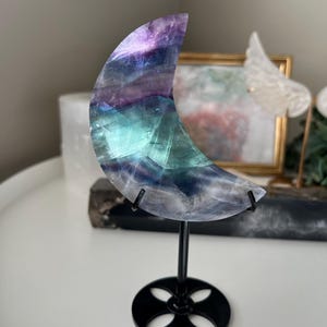 May include: A crescent moon-shaped fluorite crystal sculpture. The crystal features bands of purple, blue, and green. It is mounted on a black metal stand with a circular base. The sculpture is displayed on a white surface.