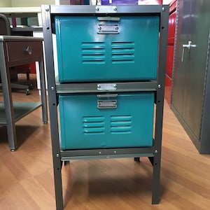 May include: Two teal metal storage drawers on a black metal frame. The drawers have a series of small holes and a metal label on the front.