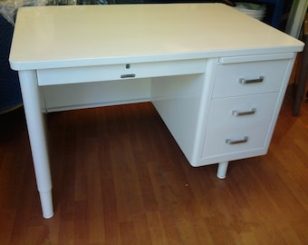 May include: A white metal desk with a rectangular top and three drawers on the right side. The desk has a single drawer on the left and silver handles. The legs are white and cylindrical.