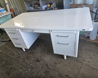 May include: A white metal desk with a glossy finish. The desk has a rectangular top, a center drawer, and two sets of drawers on either side. Each drawer has a silver handle. The desk is supported by four short legs.