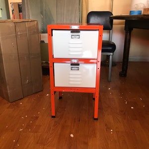May include: Two-tiered metal storage unit with orange frame and white drawers. Each drawer has a metal handle with a number on it.