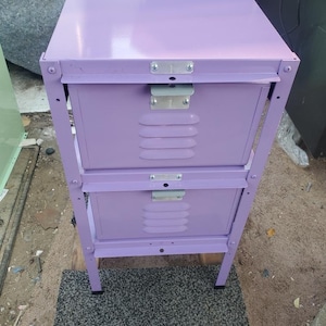 May include: A two-drawer metal locker with a purple paint finish. The locker has a metal handle and a metal latch. The locker is sitting on a gray rug.