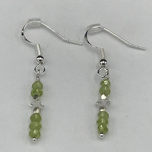 May include: A pair of dangle earrings with silver-colored hooks. Each earring features a cluster of small, light green beads, a clear, star-shaped bead, and a silver-colored bead at the bottom. The earrings are displayed against a white background.