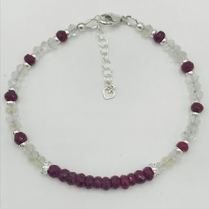 May include: A delicate bracelet featuring alternating faceted ruby and clear quartz beads. The bracelet has a silver-toned clasp and an adjustable chain with a small heart charm. The ruby beads are a deep red color.