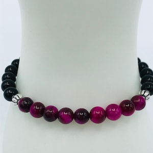 May include: A beaded bracelet featuring a combination of black and magenta-colored round beads. The bracelet has silver-colored metal accents. The beads are strung together on a flexible cord, creating a piece of jewelry.