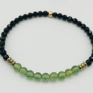 May include: A beaded bracelet featuring black and green beads with gold accents. The bracelet has a central row of green beads, flanked by black beads and gold-colored spacers. A small gold bead is also present.