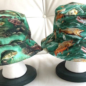 May include: Two bucket hats with a green background and a print of fish. The hat on the left has a print of bass fish, and the hat on the right has a print of trout fish. The hats are on display on a white mannequin head.