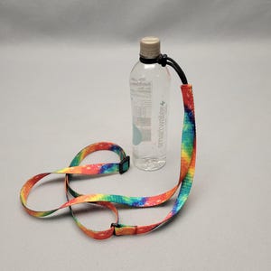 May include: A clear plastic water bottle with a black cap and a colorful tie-dye lanyard. The lanyard is made of a woven material with a rainbow pattern and a black plastic buckle.