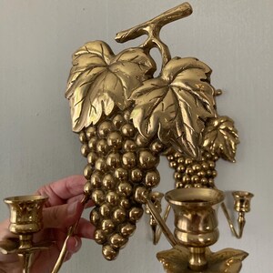 May include: Gold-colored wall sconces featuring a grape cluster design. The sconces have detailed leaves and grape clusters, with candle holders. The sconces are made of metal and are designed to hold candles.
