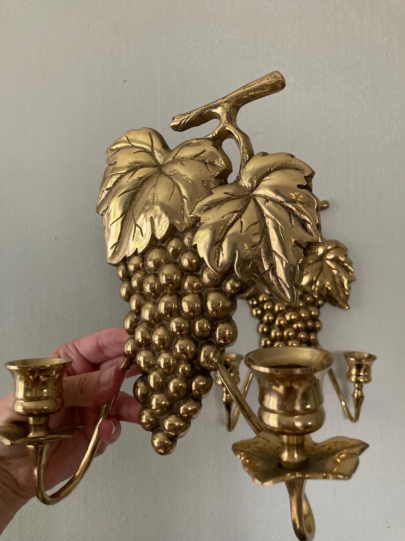 May include: Gold-colored wall sconces featuring a grape cluster design. The sconces have detailed leaves and grape clusters, with candle holders. The sconces are made of metal and are designed to hold candles.