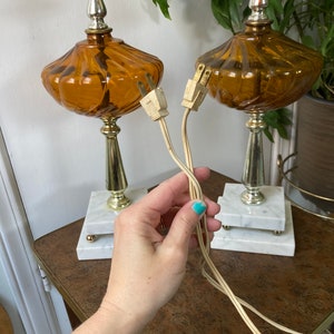 May include: Two amber glass table lamps with white marble bases and gold accents. The lamps have a fluted design and are plugged in with white cords.
