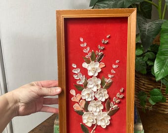 May include: A framed floral art piece featuring white flowers and green leaves against a red background. The artwork is encased in a wooden frame. The piece is held by a person's hand.