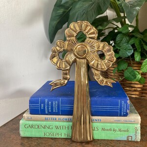 May include: A gold-colored metal decorative bow, possibly a door knocker or wall hanging, stands upright on a stack of vintage books. The bow has intricate detailing and a vertical stem. The books are blue and green, with titles visible.