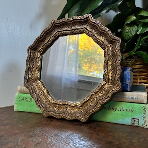 May include: A round mirror with a decorative, scalloped, antique gold-toned frame featuring intricate embossed patterns. The mirror is positioned upright, resting against a stack of two books, one of which is green and titled Garden Month by Month.