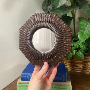 May include: A small, round mirror with a dark brown, textured frame. The frame has a woven or braided appearance. The mirror is held in front of a stack of books. The background includes green plants and a wicker basket.