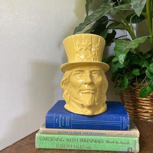 May include: A yellow ceramic Uncle Sam head planter featuring a tall top hat adorned with embossed stars. The planter is resting on a stack of three books titled The Deepening Stream, Better Flower Arrangements, and Gardening with Perennials.