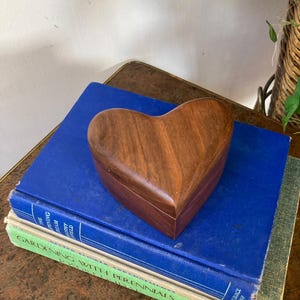 May include: A heart-shaped wooden box with a rich, brown finish sits atop two vintage books. The box is made of a dark wood with visible grain. The books are blue and green, with gold lettering. The scene is set on a brown surface.