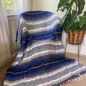 May include: A blue, white, and gray woven blanket with a geometric pattern draped over a chair. The blanket has a fringed edge and is made of a soft, textured material. The blanket is approximately 70 inches long and 50 inches wide.