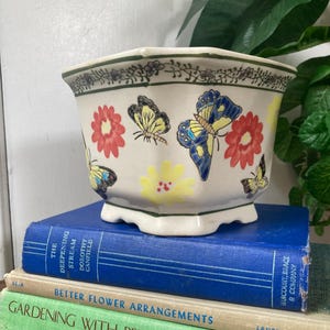May include: A cream-colored ceramic planter with a scalloped rim, decorated with red, yellow, and blue butterflies and flowers. The planter sits atop a stack of vintage books with titles like "The Deepening Stream" and "Gardening with Perennials."