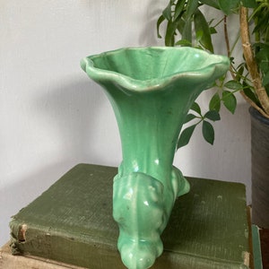 May include: A green ceramic vase with a trumpet-shaped opening and a sculpted base. The vase is sitting on a stack of books.